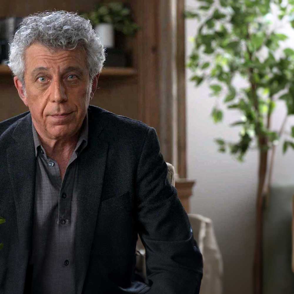 Eric Bogosian as Daniel Molloy in Interview with the Vampire. He sits in a chair in a conference room set, looking at the camera to deliver his 