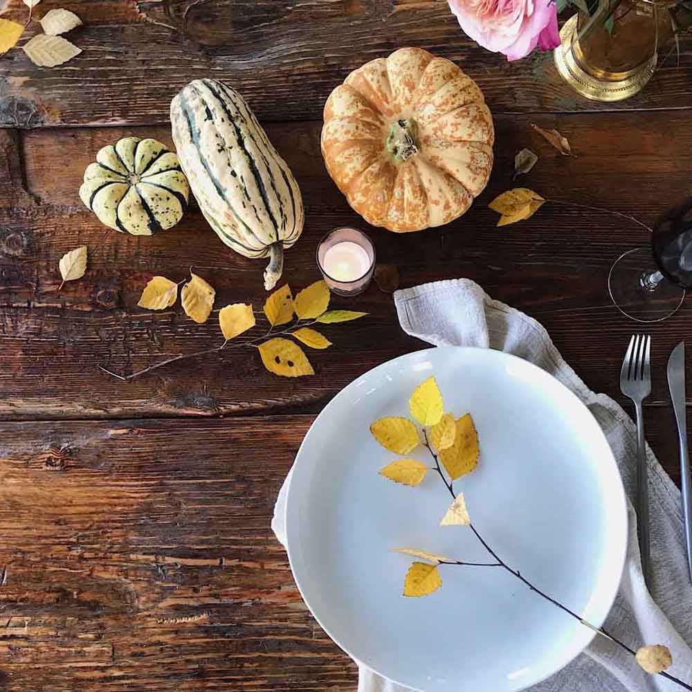 An overhead view of a rough-hewn wooden table decorated for autumn with acorn and delicate squashes, a small pumpkin, a ceramic plate, linen napkin, and branches of autumn leaves.