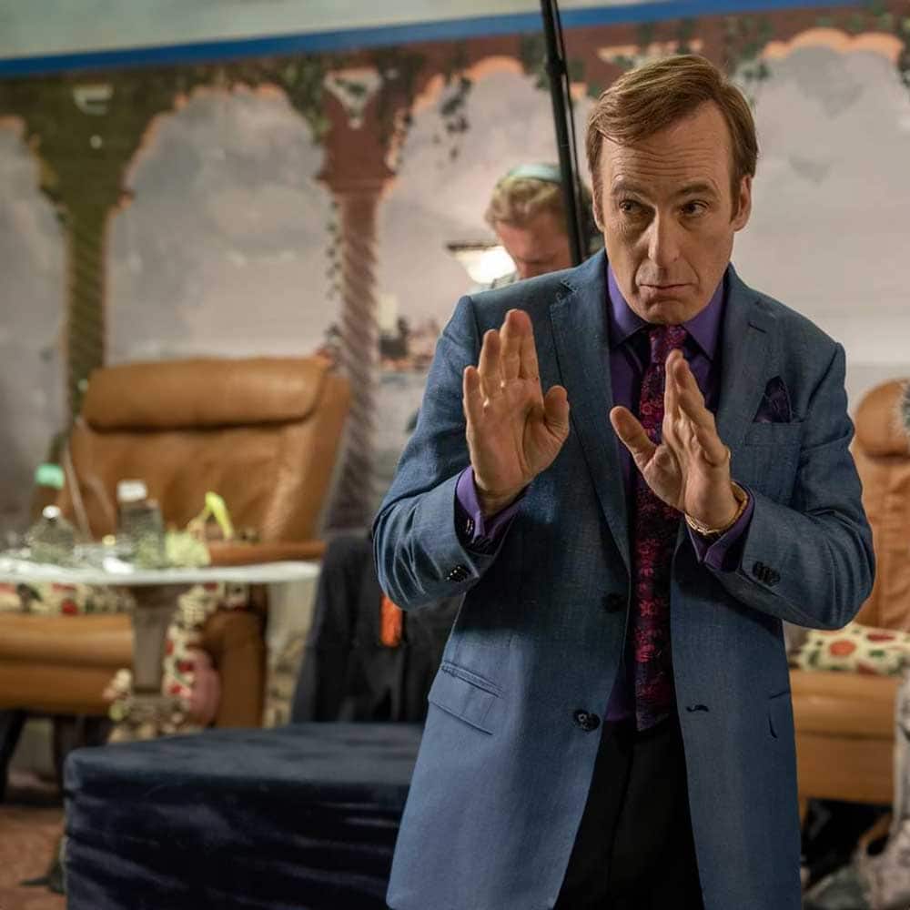 Bob Odenkirk as Saul Goodman, dressed in a blue suit with a purple shirt, directs a video, giving instructions to someone offscreen. Behind him, we can see a boom operator and the wall of a salon.