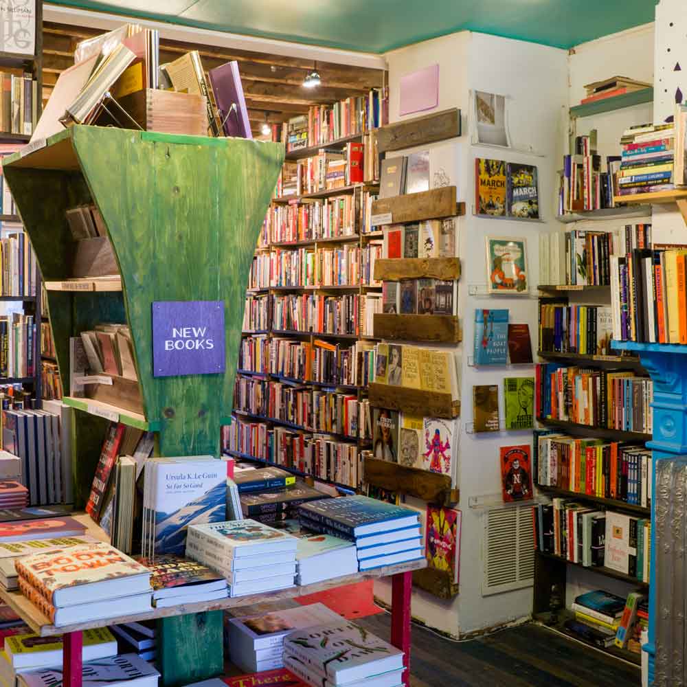 A bookshop: in the foreground is a table with new books, in the background are floor-to-ceiling shelves of used books, and to the right is a face-out display.