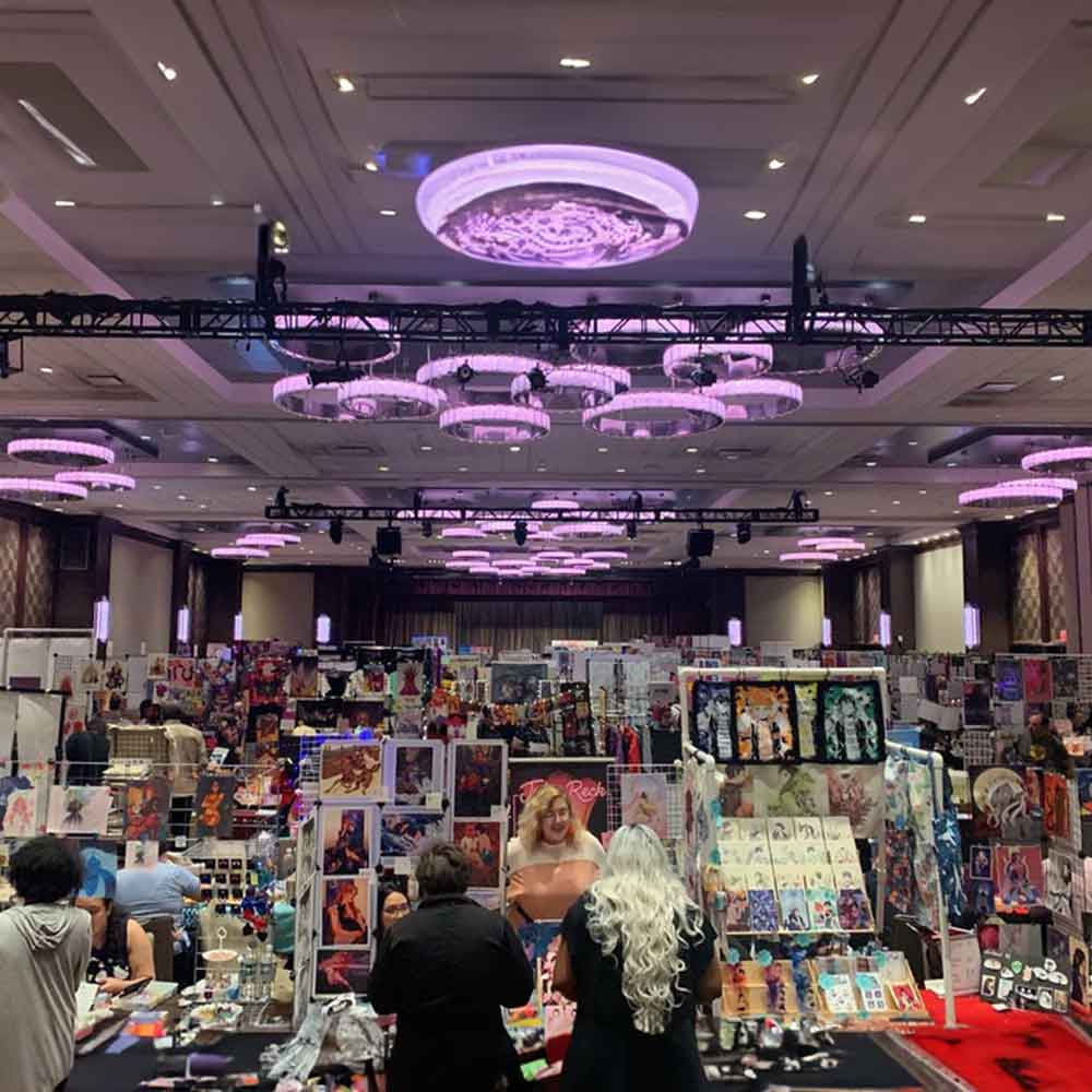 The inside of a convention hall. In the foreground, attendees talk with artists at their tables in the artist alley, each dense with prints and other wares on display. In the background, we nee more tables stretching into the distance.