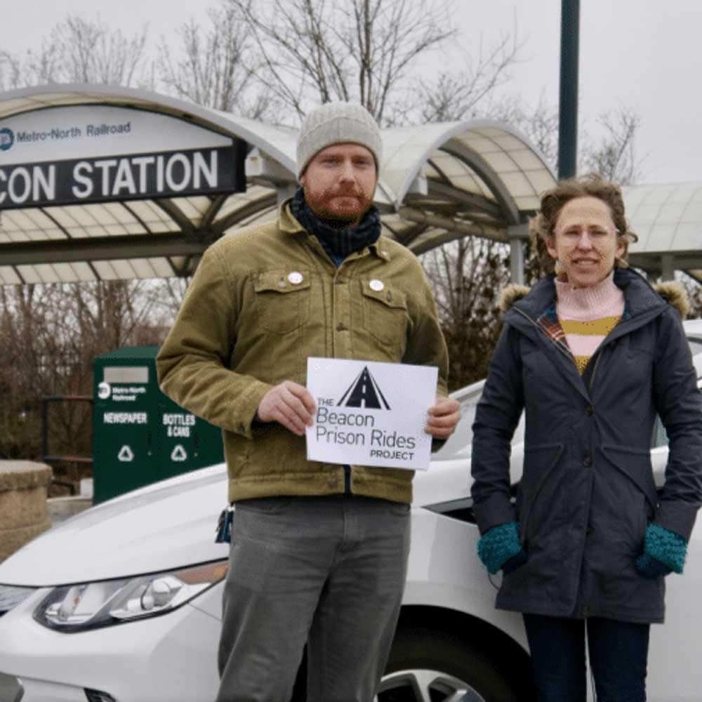 A man and a woman, each dressed for chilly weather, stand next to a car outside the Beacon Metro North Train Station. The man holds a sign that reads 
