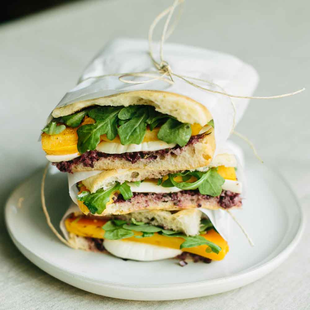Three stacked sandwiches on a white ceramic plate. Each sandwich contains tomato, arugula, fresh mozzarella, and homemade spread, and is banded with white deli paper and tied with cotton twine. 